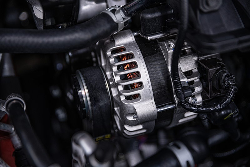 Close-up of a car engine alternator with a drive belt, showing copper wiring through its vented metal casing.