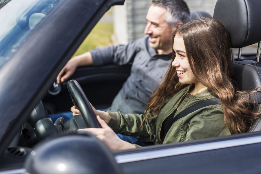 A smiling teenage girl drives a convertible with her father in the passenger seat.