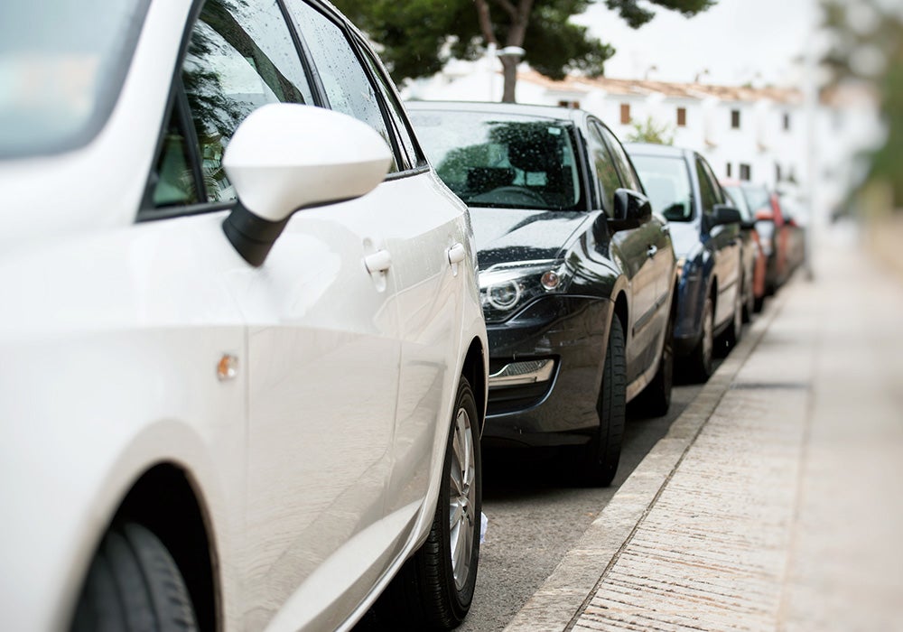 A line of cars parked parallel along a street curb next to a sidewalk.